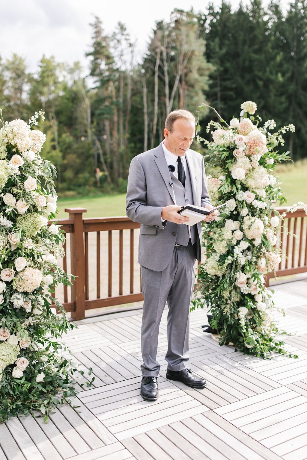 A man in a gray suit reading a book or speech at an outdoor wedding ceremony with floral arrangements on either side, set on a wooden platform with a green forest background.