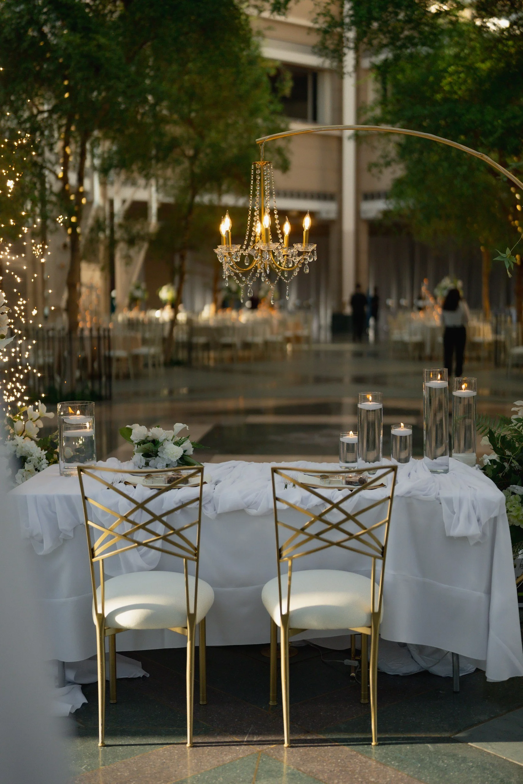 Elegant outdoor wedding reception setup with a table, gold chairs, floral arrangements, candles in glass holders, and a chandelier hanging from an overhead branch, surrounded by trees and string lights.