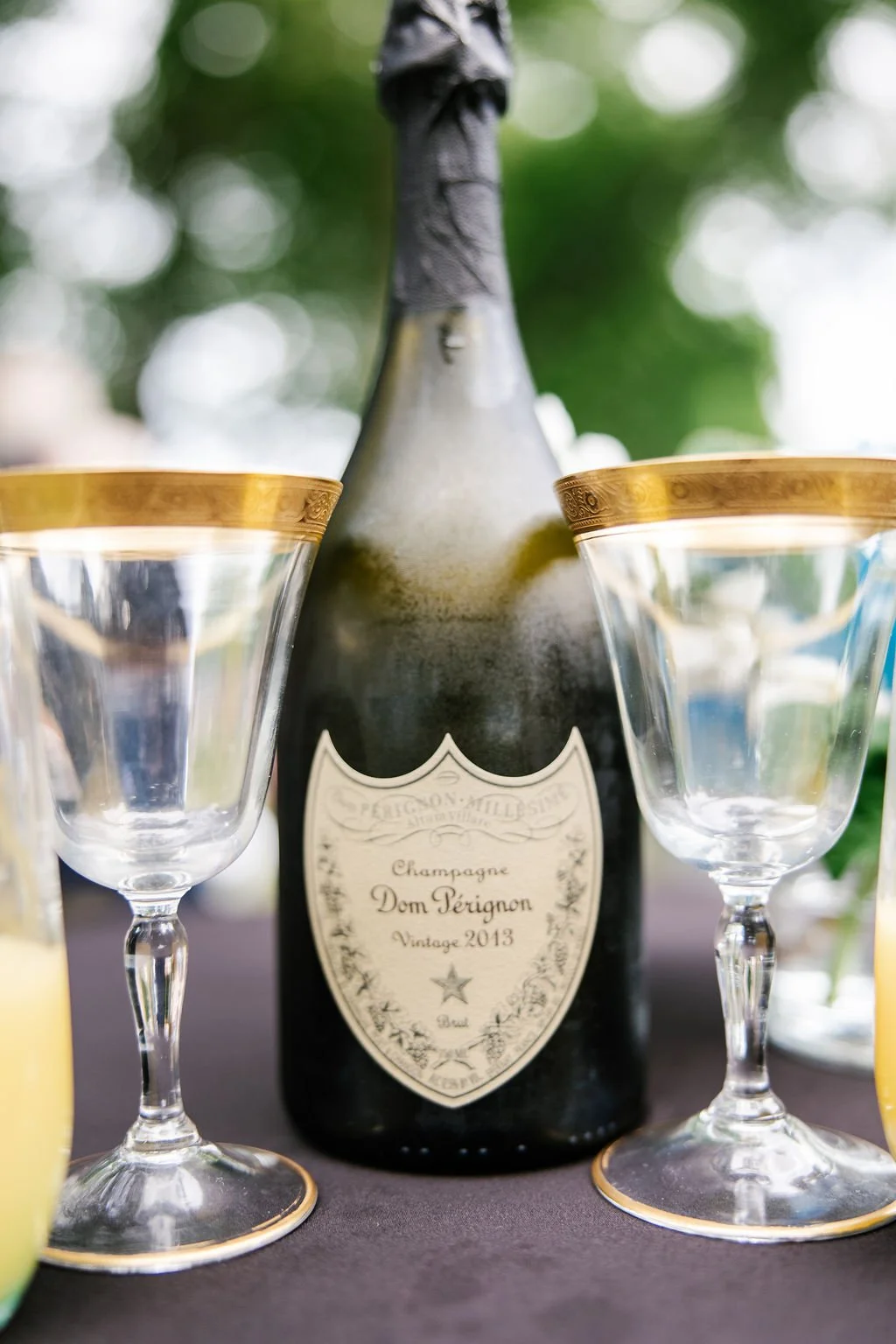 A bottle of Dom Pérignon vintage 2013 champagne between two empty champagne glasses with gold rims, on a dark table with greenery in the blurred background.