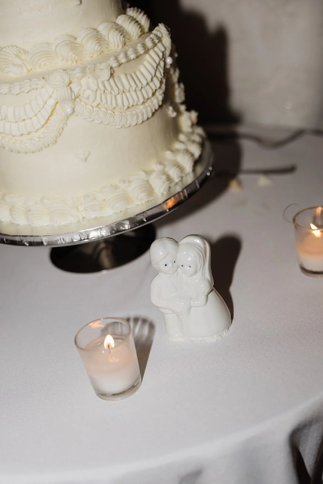 A wedding cake decorated with white frosting and pearl accents, placed on a table. In front of the cake, there is a ceramic figurine of a bride and groom, flanked by small lit candles in glass holders.
