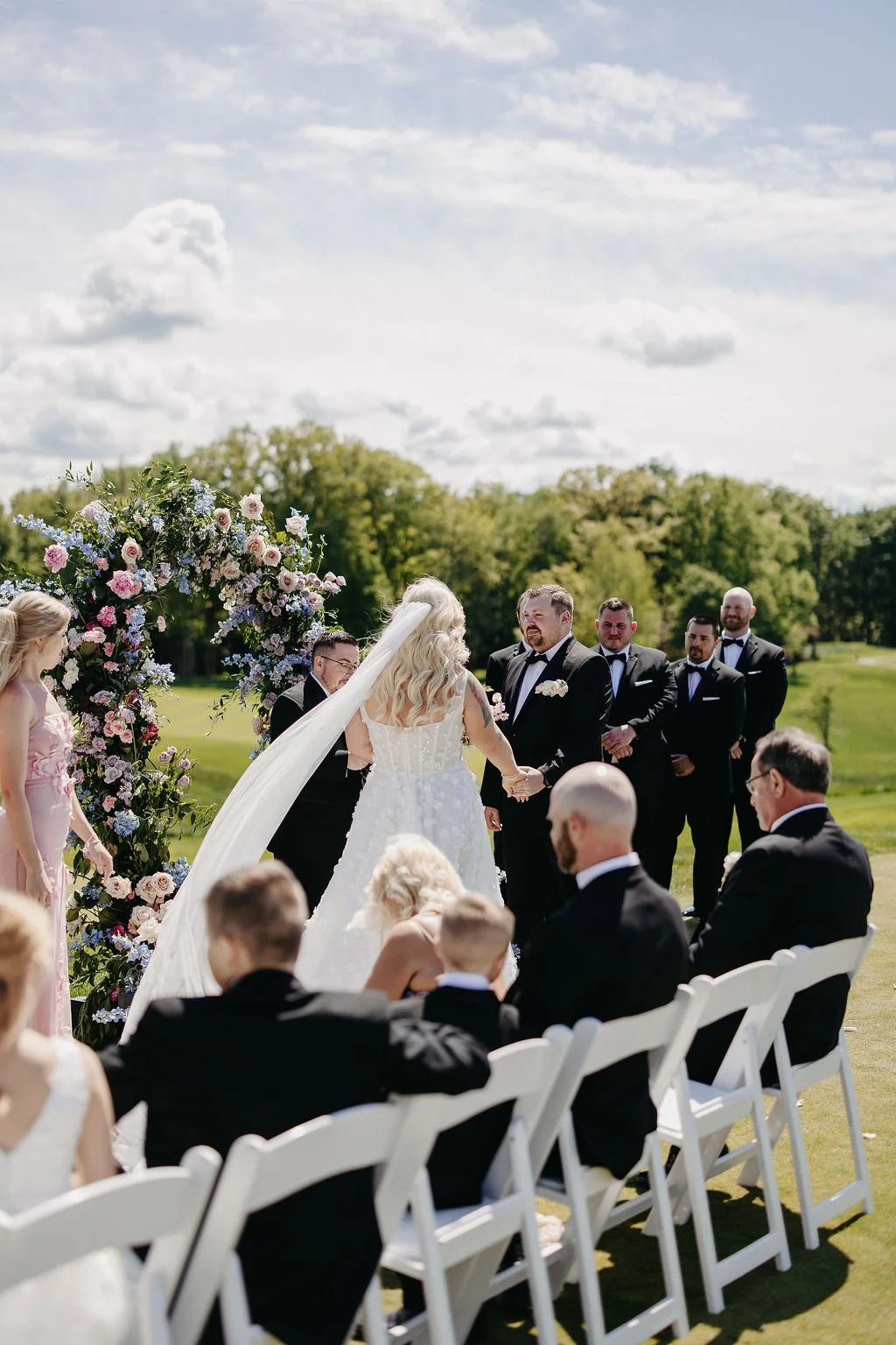 A wedding ceremony taking place outdoors with the bride and groom holding hands in front of an officiant, surrounded by bridesmaids and groomsmen, with guests seated on white chairs, and a floral arch in the background under a partly cloudy sky.