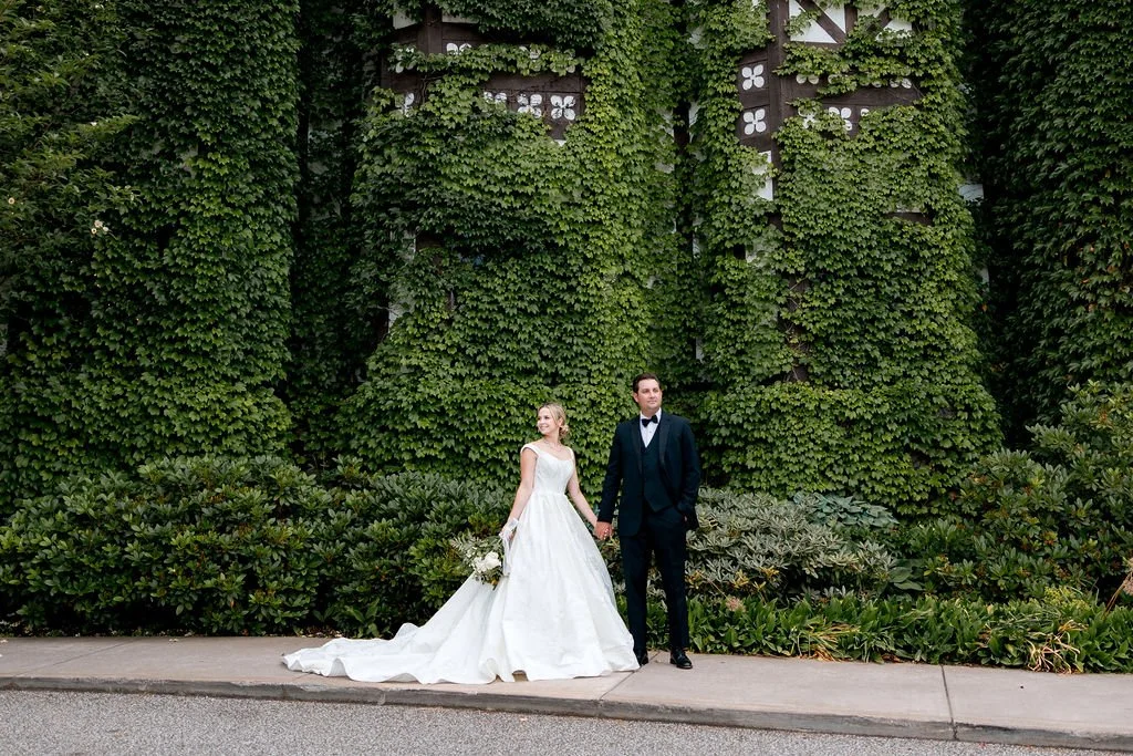 Bride in a white wedding gown holding a bouquet, groom in a black tuxedo with a bow tie, standing outdoors on a sidewalk with lush green ivy-covered wall behind them.