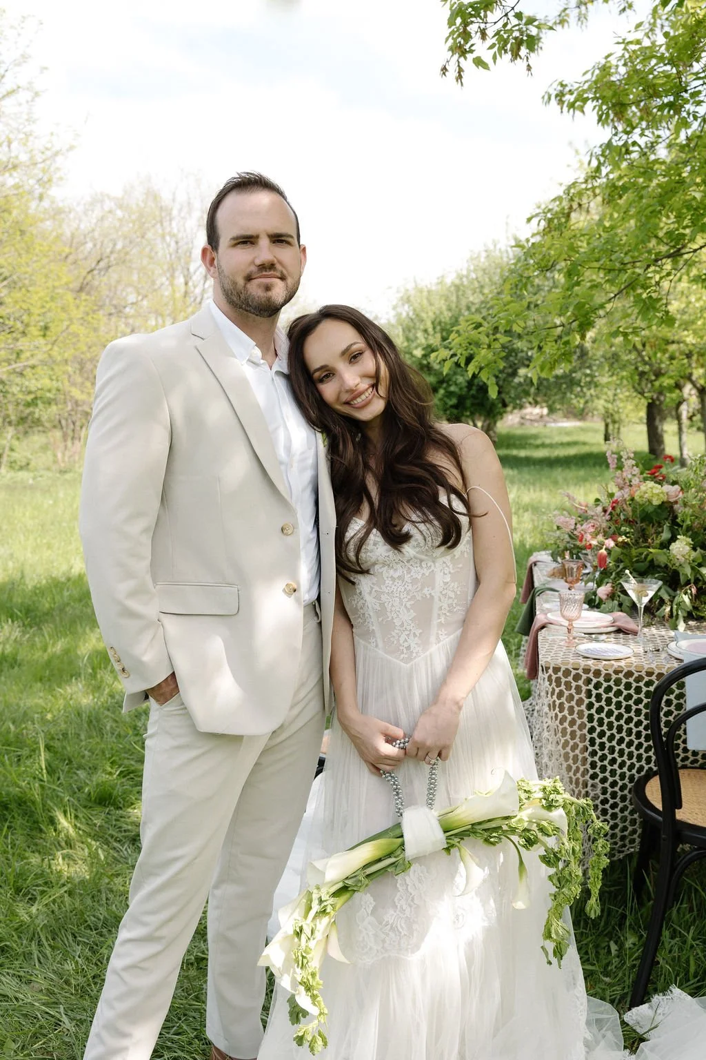 A couple stands together outdoors at a wedding reception, with the woman holding a bouquet of white flowers.