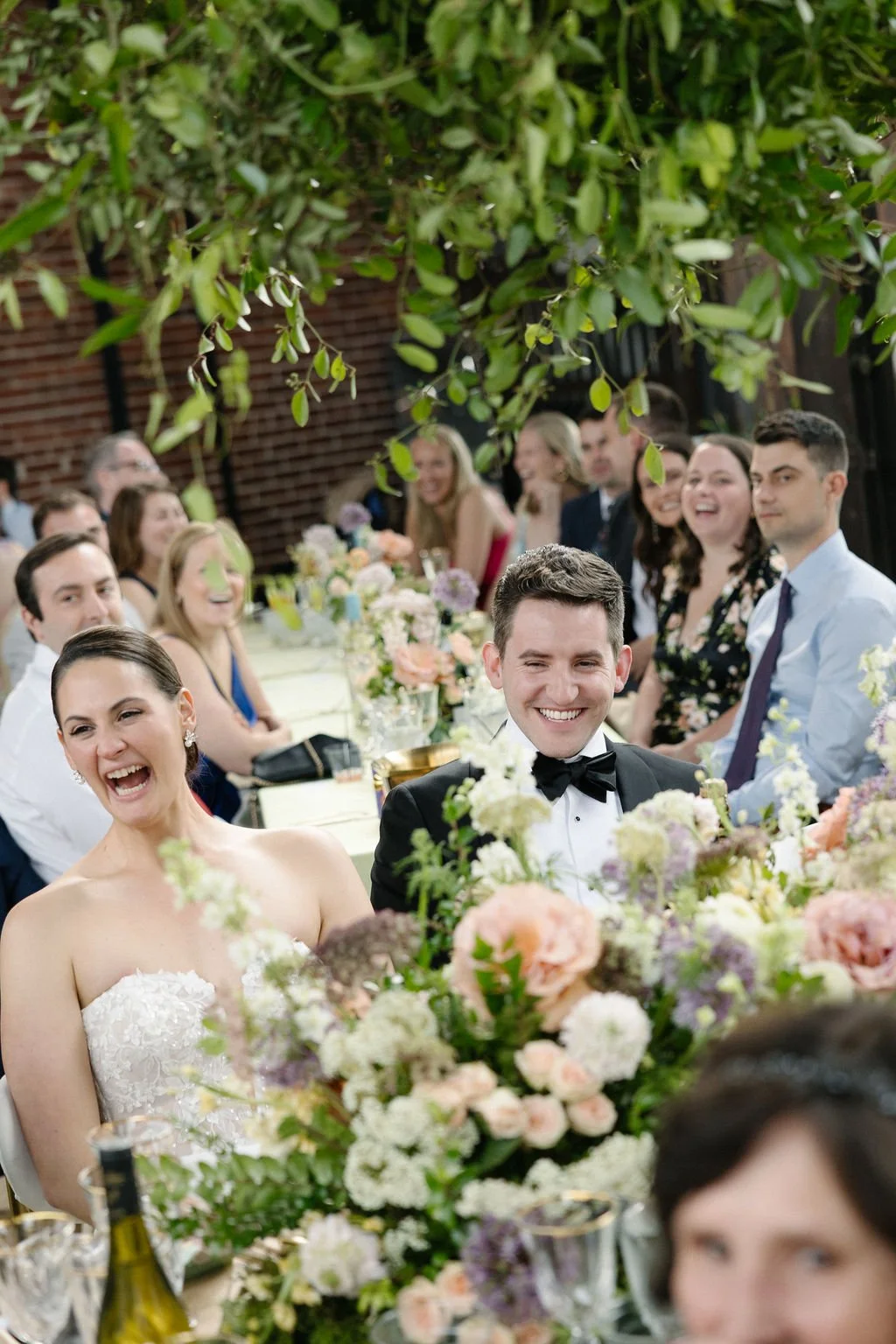 Wedding celebration with smiling bride and groom sitting at a decorated table surrounded by friends and family at an indoor venue with greenery and floral arrangements.
