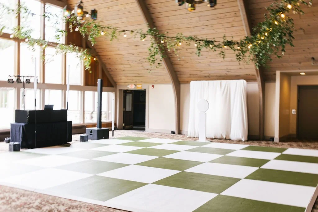 Empty event space with a checkered green and white dance floor, decorated with greenery and string lights hanging from the ceiling, with large windows on one side and a DJ booth on the other.