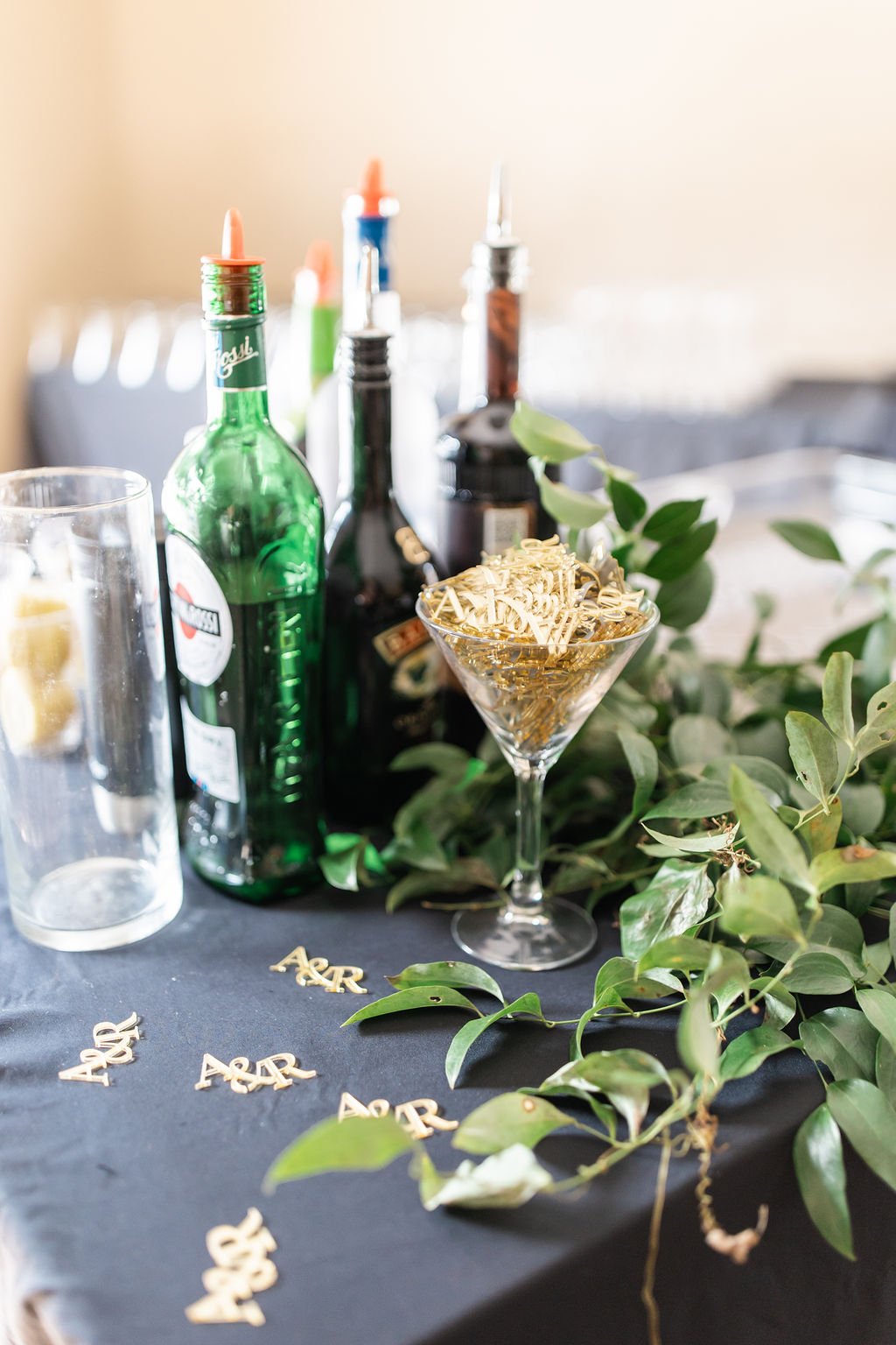 Party table with greenery, gold confetti, a martini glass filled with gold ribbon, and bottles of alcohol including gin, vodka, and whiskey.