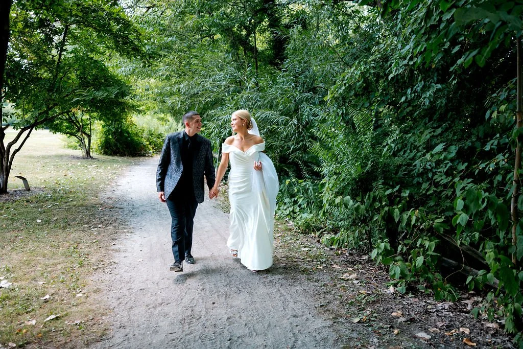 A couple, dressed in wedding attire, walking hand in hand on a dirt path through a lush, green forest.