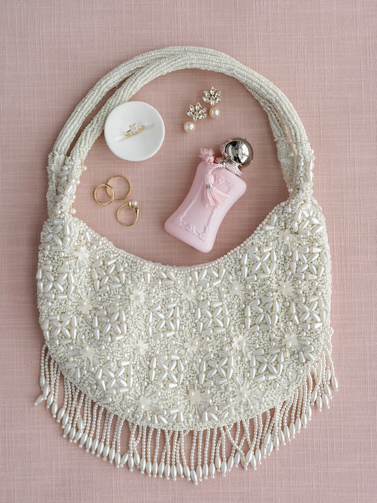A white beaded handbag decorated with pearls and flowers, placed on a pink fabric background, with jewelry including pearl and gold earrings, a ring, a diamond ring, a pink decorative perfume bottle, and pearl earrings placed on or near the handbag.