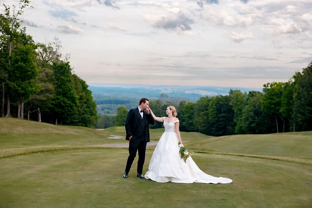 A bride and groom in wedding attire on a golf course, with the groom touching the bride's face and the bride holding a bouquet, surrounded by trees and a cloudy sky.