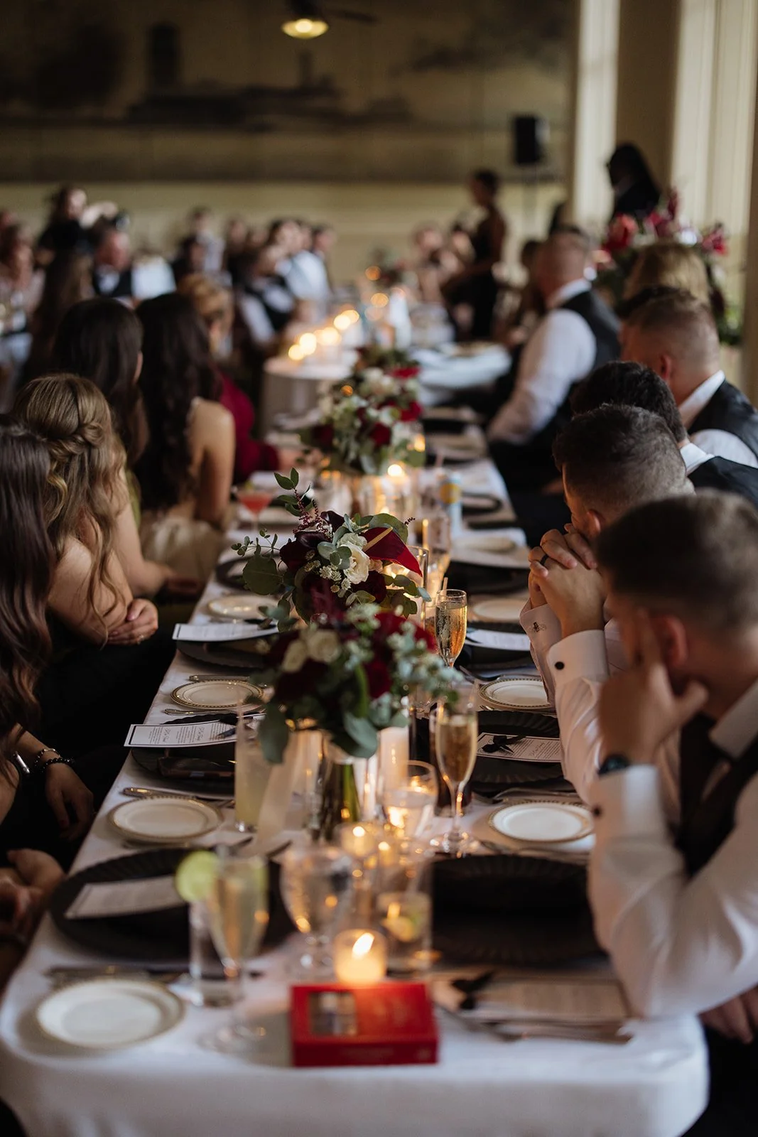 A formal dinner event with a long table adorned with floral centerpieces and candlelight, surrounded by guests dressed in evening attire, possibly at a wedding or celebration.