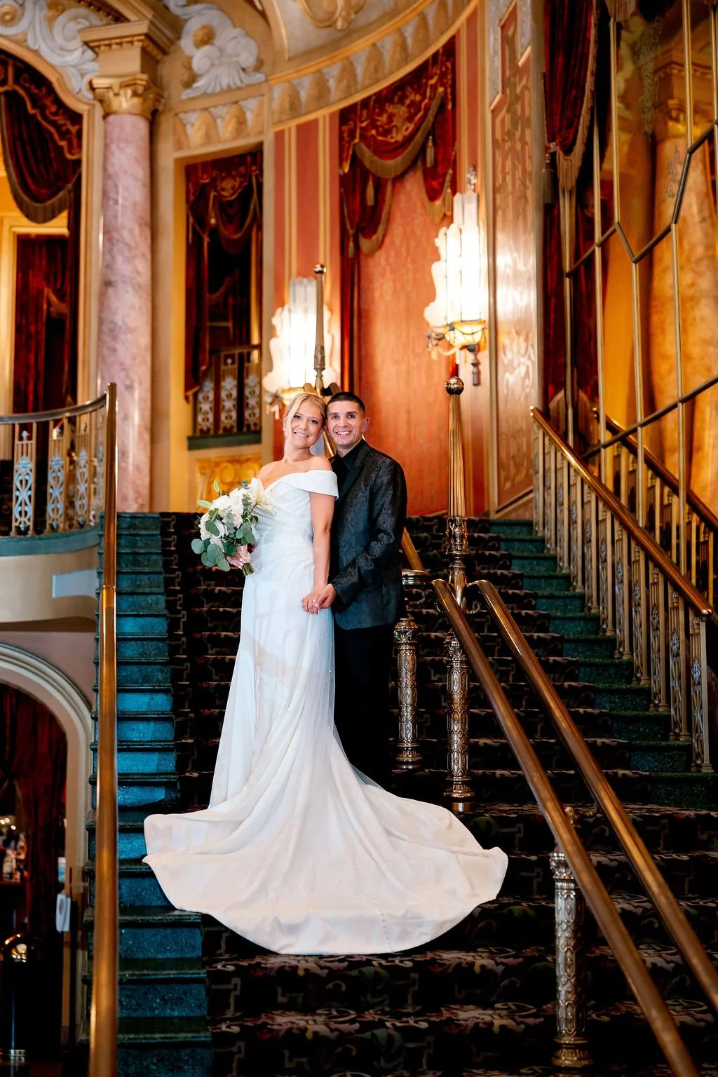 A bride and groom standing together on a grand staircase inside an ornate, historic building, smiling and holding hands. The bride is in a white wedding gown holding a bouquet of flowers, and the groom is dressed in a black tuxedo.