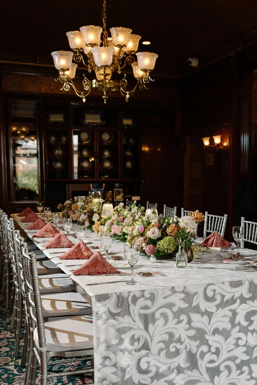 A formal dining room with a long table set for a meal, decorated with a floral centerpiece, pink napkins, and glassware, illuminated by a chandelier above.