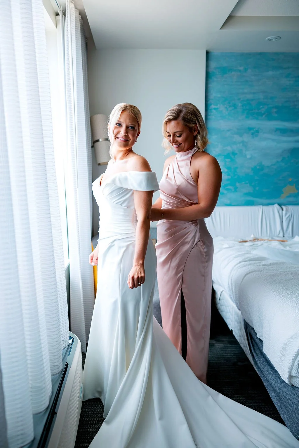 A bride in a white wedding dress smiling while a woman, possibly her bridesmaid or maid of honor, helps her adjust her gown in a hotel room with natural light coming through the window.