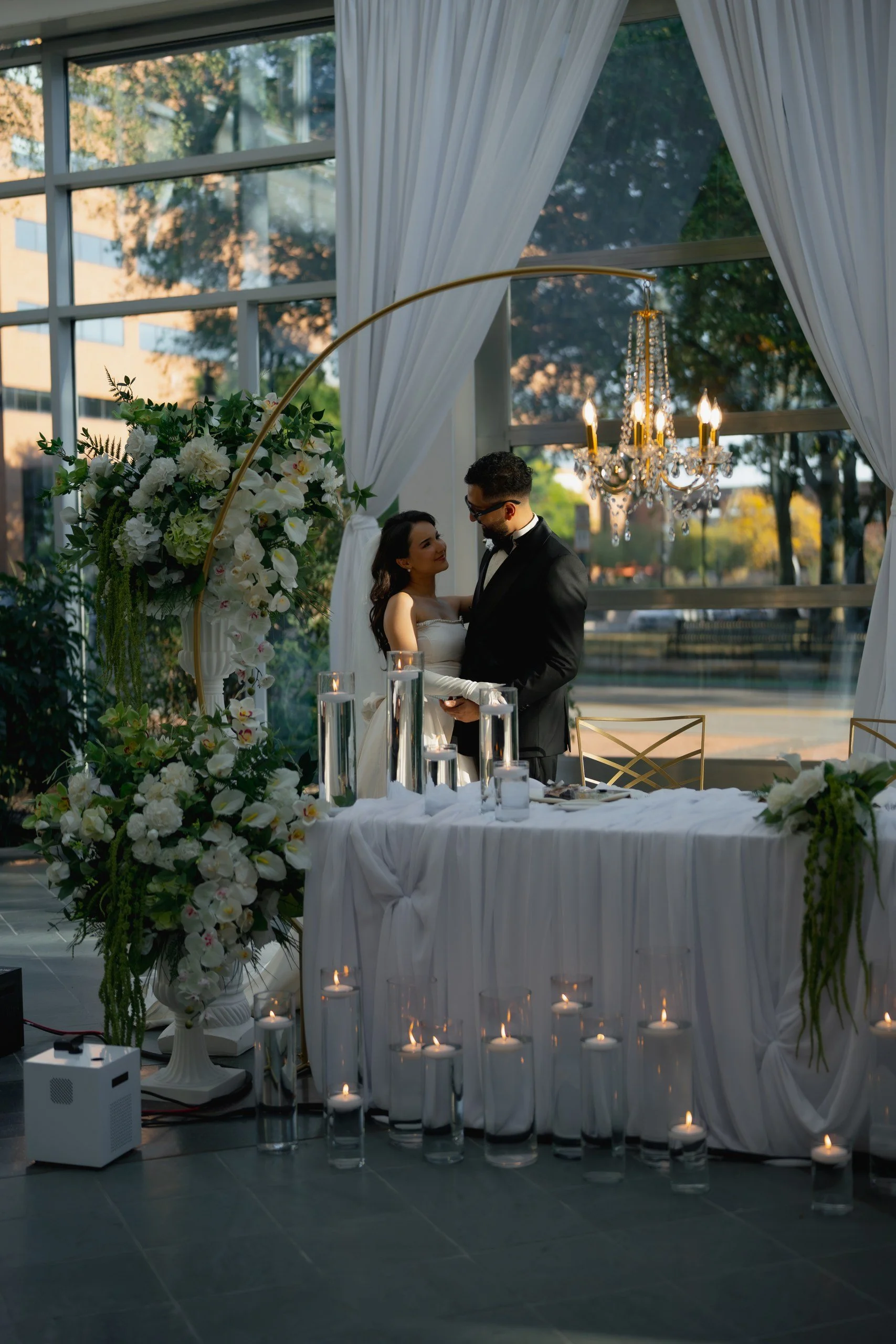 A wedding ceremony setup inside a glass-walled venue with white drapery, a chandelier, and floral arrangements, with a bride and groom holding hands.