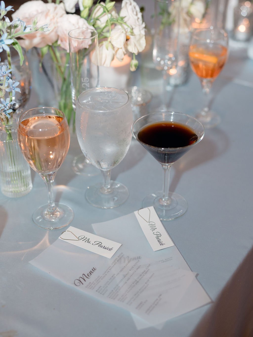 Elegant table setting with wine glasses filled with rosé, water, and red wine, surrounded by floral arrangements and a printed menu.