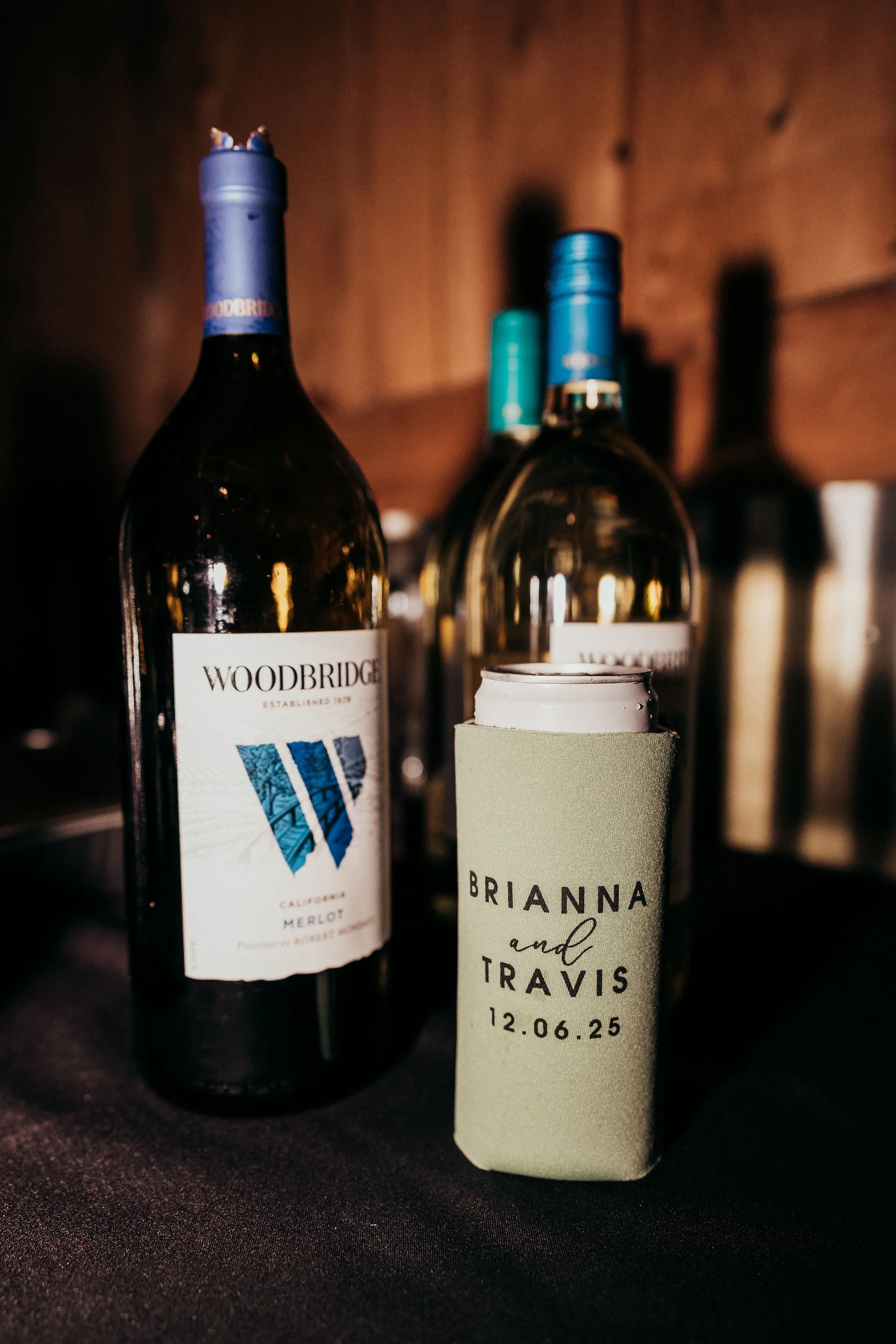 Close-up of wine bottles and a personalized wedding favor with names Brianna and Travis and the date 12.06.25, on a dark surface with a wooden background.