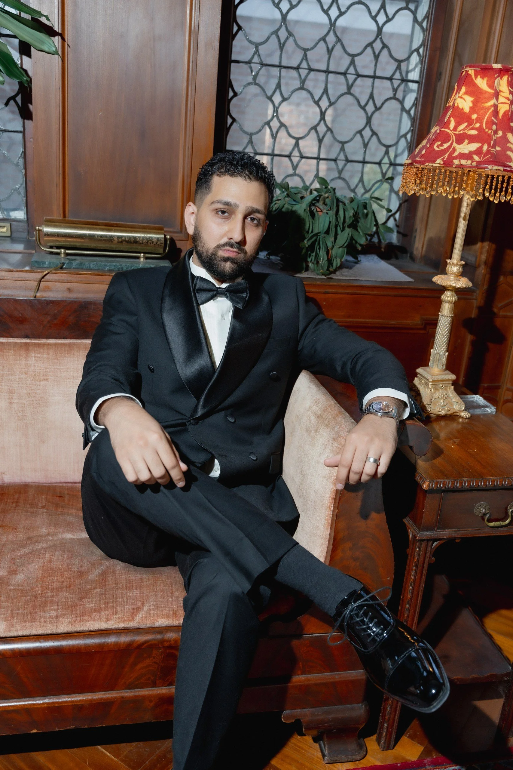 A well-dressed man in a tuxedo with a bow tie is sitting on a vintage wooden sofa in a room with wooden paneling, a window with decorative ironwork, a potted plant, an ornate table lamp, and vintage decor.