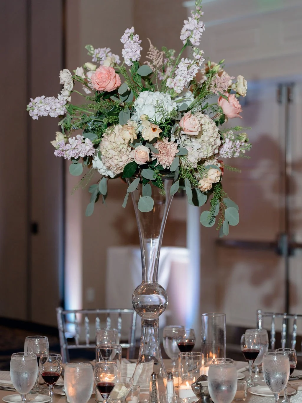 Elegant floral centerpiece with pink and white flowers in a tall glass vase on a table set with water glasses, wine glasses, and candles, at a formal event.