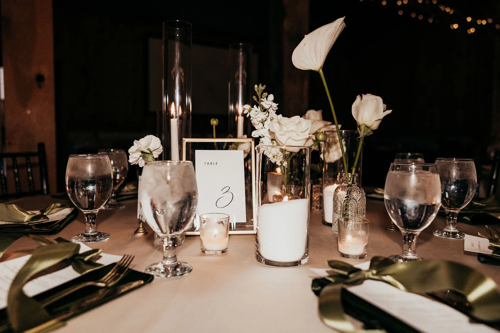 A decorated table with white flowers in vases, candles, and glassware in a dimly lit setting, likely for a formal event or dinner.