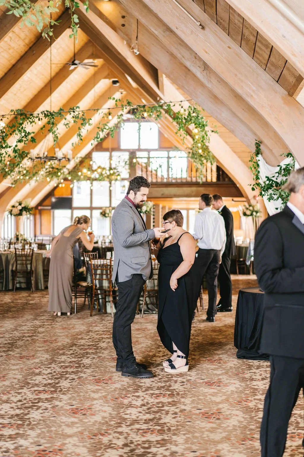 People socializing at a formal event in a decorated wooden venue with high vaulted ceilings, hanging greenery, and large windows.