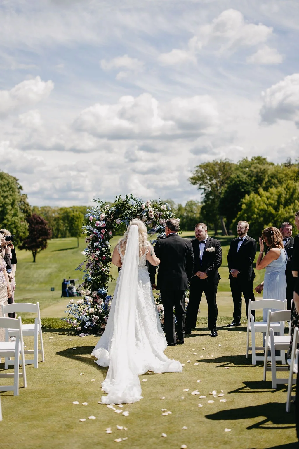A wedding ceremony outdoors with a bride and groom facing an officiant, surrounded by guests, under a floral arch on a sunny day with a partly cloudy sky and green trees in the background.
