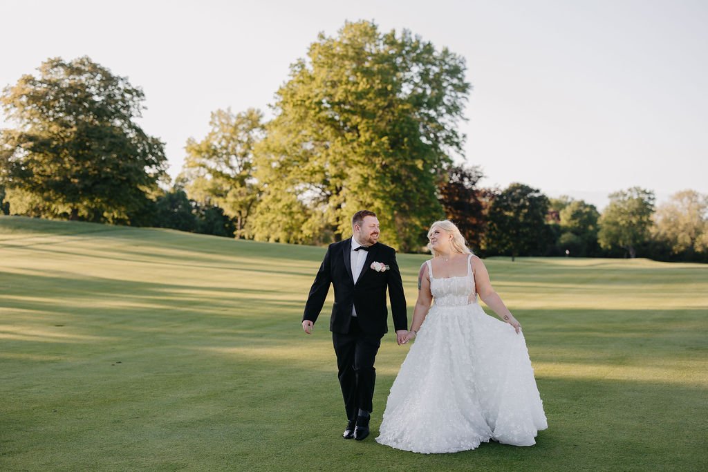 A newlywed couple walking hand in hand on a golf course, surrounded by green trees and grass, enjoying a sunny day.