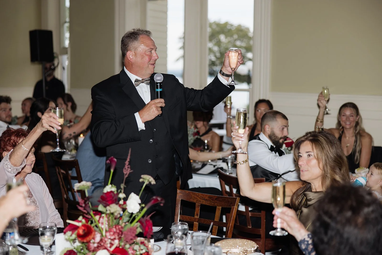 A man in a tuxedo holding a microphone in one hand and a champagne flute in the other, giving a toast at a formal dinner event. Guests in the background are also raising their glasses.