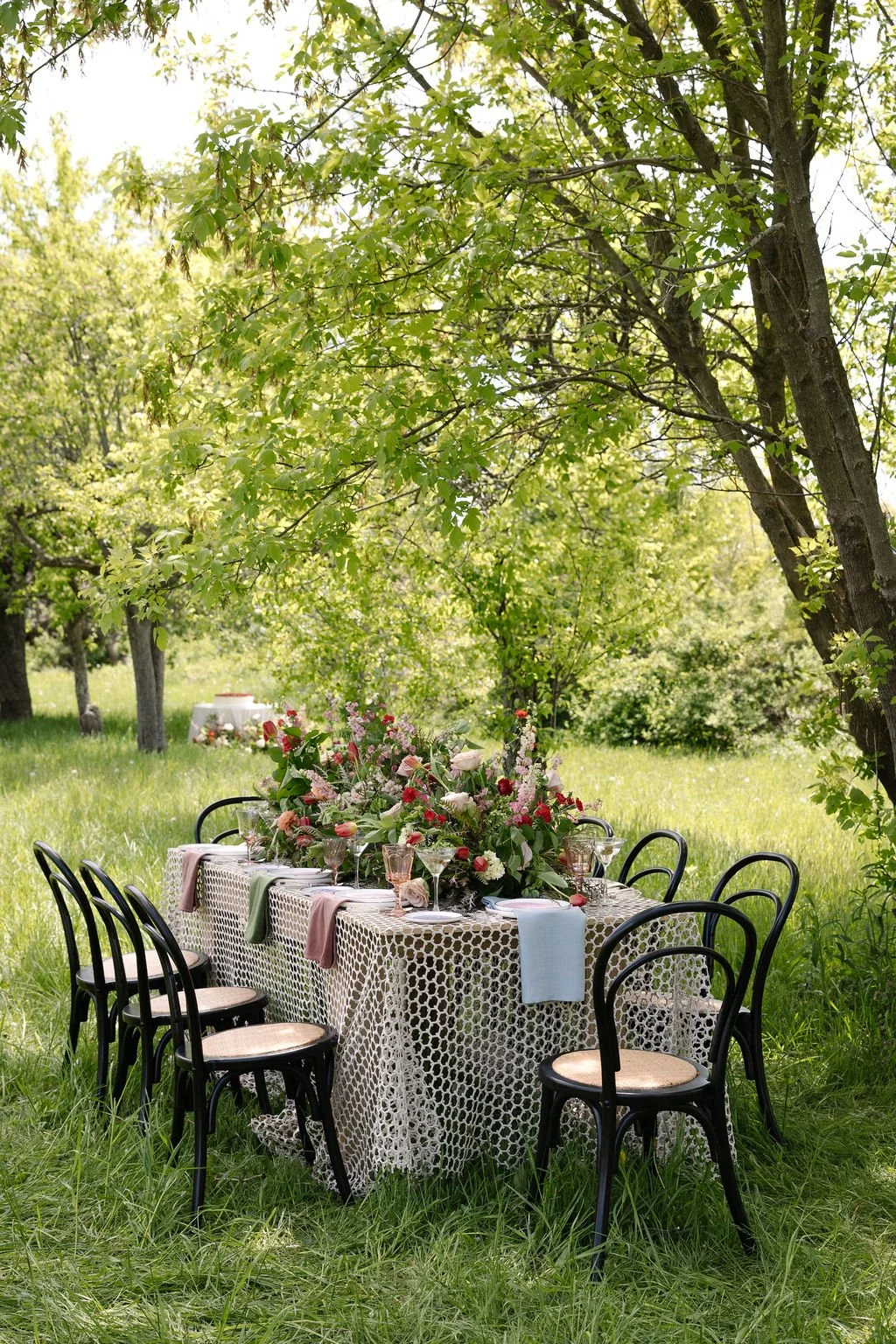 A table set for an outdoor meal in a lush green garden, decorated with a large floral centerpiece, surrounded by black chairs with light-colored seats, under shade of green trees.