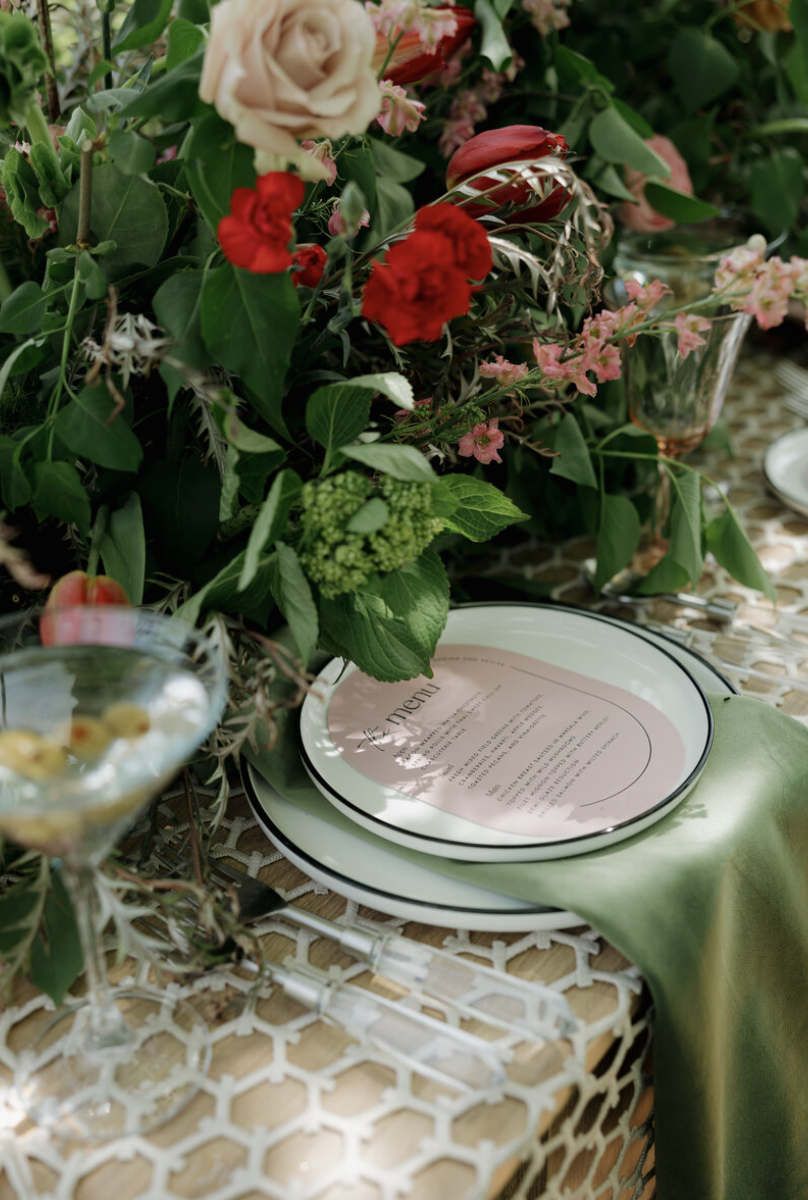 A table setting with a floral centerpiece, plates with a menu, a green napkin, and glassware on a wicker table.