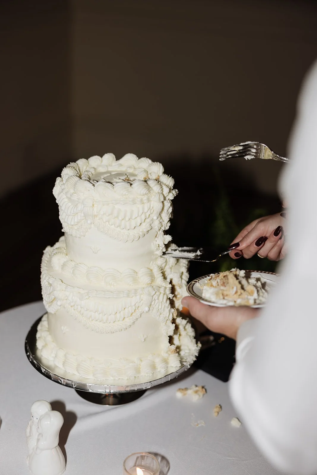 A person serving a slice of a large white wedding or special occasion cake with intricate piping and decoration, on a table.