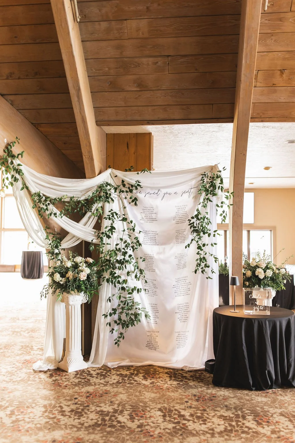 Wedding seating chart displayed on a white fabric backdrop decorated with green foliage, flanked by flower arrangements in vases, with a small table holding a transparent box labeled for cards and gifts, set inside a wood-beamed room.