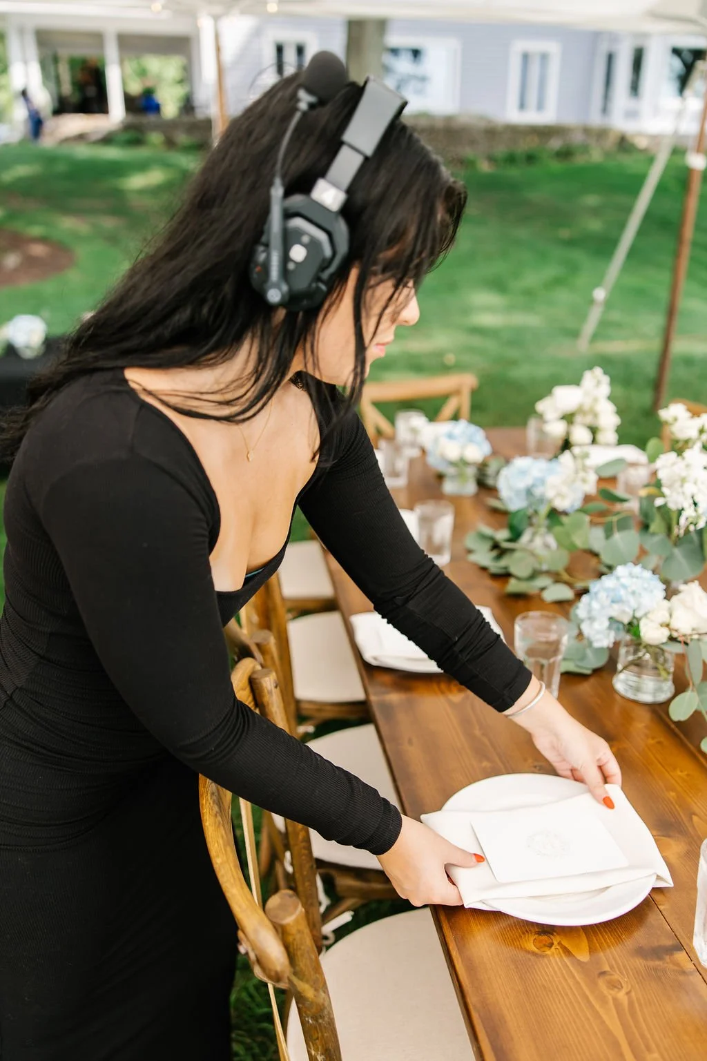 A woman with long dark hair, wearing a black dress and headphones, is setting a table with a white plate and napkin at an outdoor event decorated with flowers and greenery.