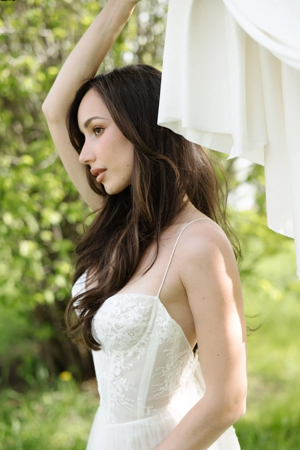 A young woman with long brown hair in a white dress standing outdoors near white curtains and green foliage.