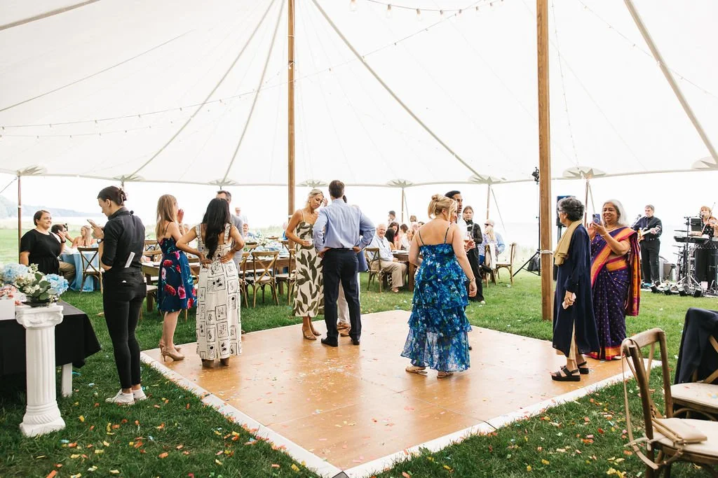People dancing and socializing under a large outdoor tent at a celebration or wedding reception.