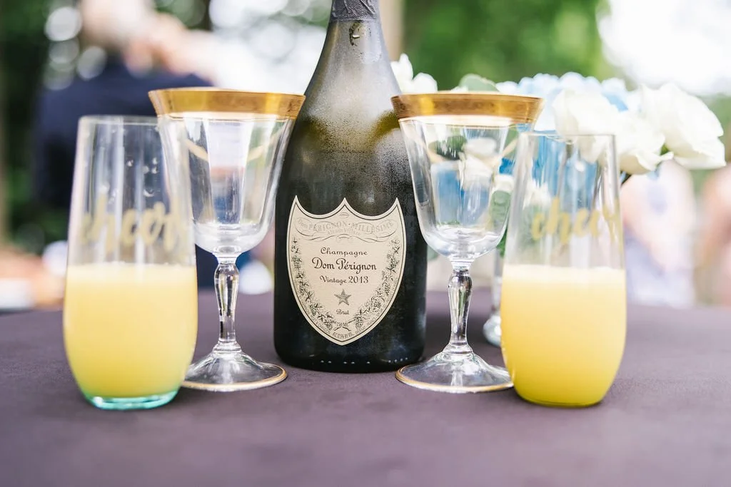 A bottle of Dom Pérignon champagne with four glasses, two filled with yellow liquid, on a table outdoors with a blurred background of people and greenery.