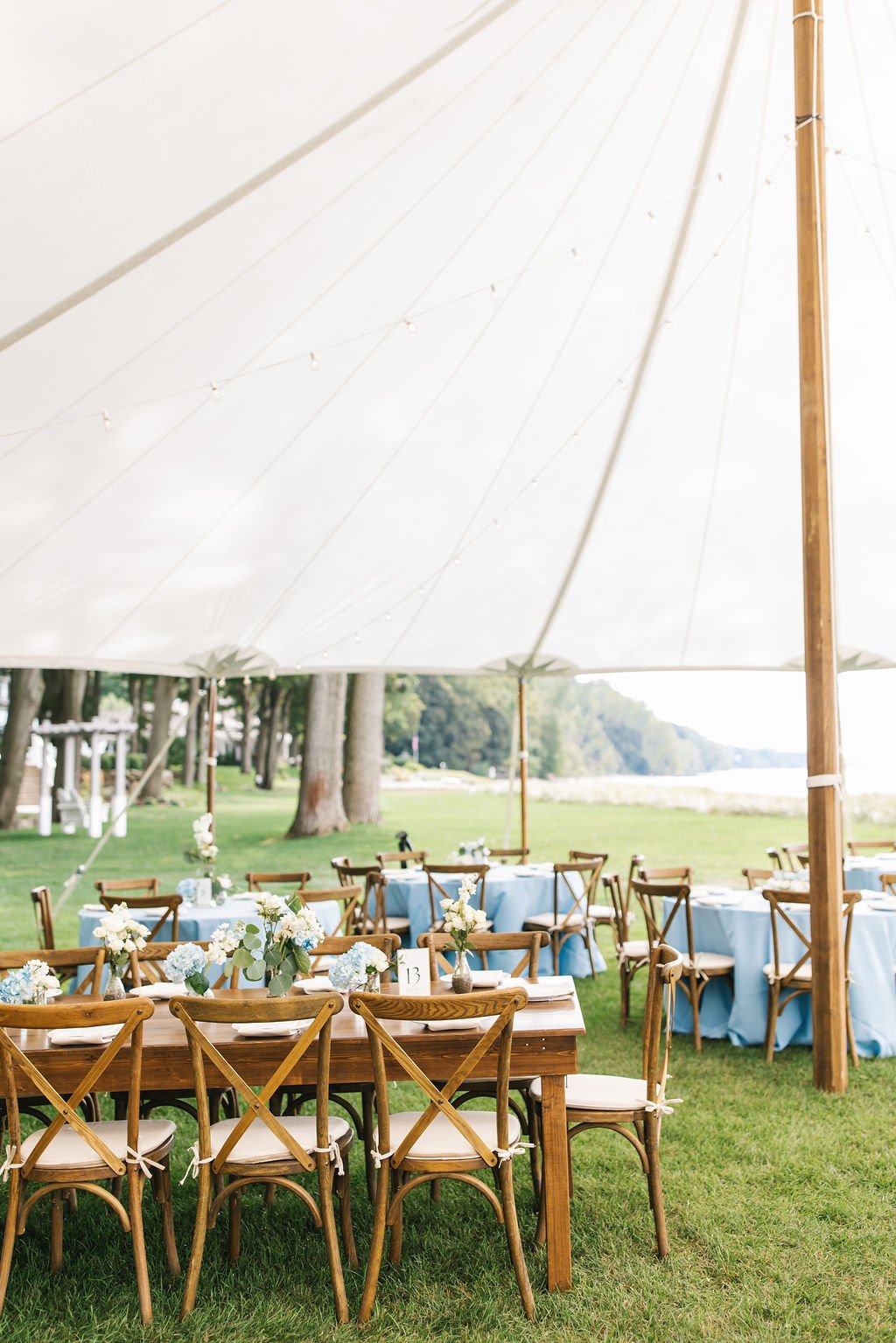 Outdoor wedding reception setup with wooden tables and chairs, decorated with white flowers and greenery, under a large white canopy on a grassy area near water and trees.