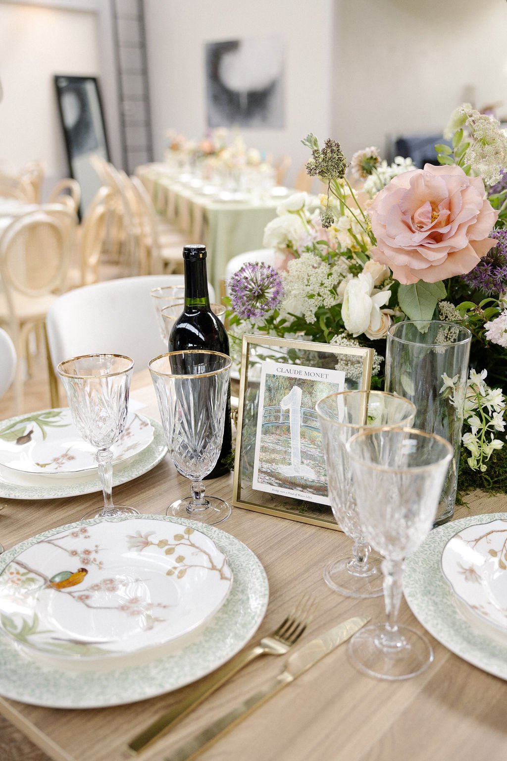 Table set for a formal event with fine china, crystal glasses, a bottle of wine, and a floral centerpiece with pink roses, white flowers, and greenery.