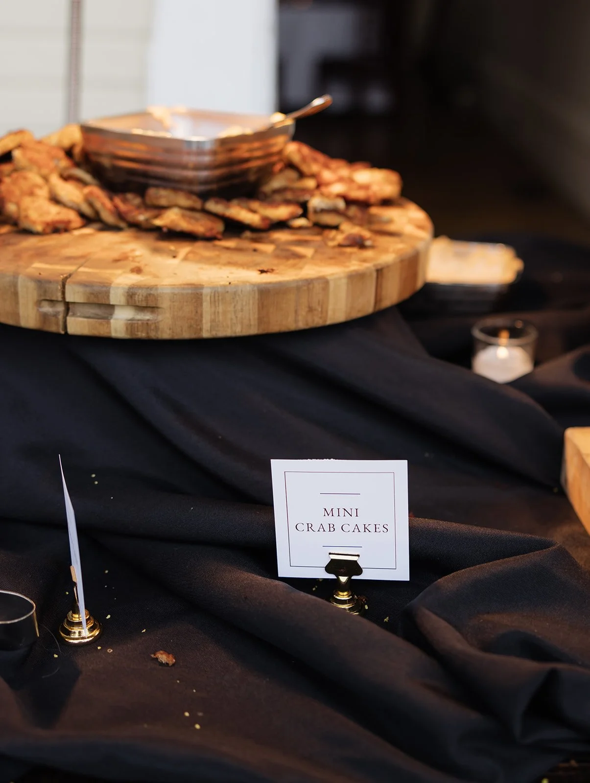 A display of mini crab cakes on a round wooden cutting board, with a small white sign that reads 'Mini Crab Cakes' placed in front of the food, set on a black cloth tablecloth.