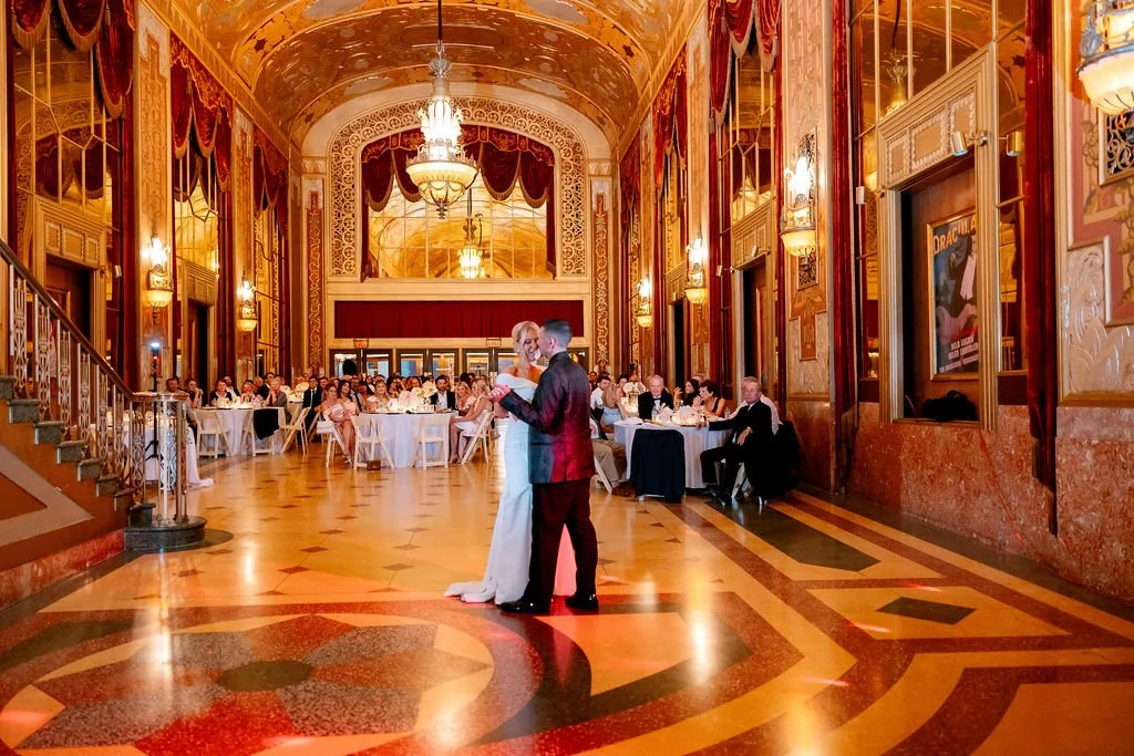 A couple dances in the center of a grand, ornate ballroom filled with seated guests at tables, during a wedding reception or special event.
