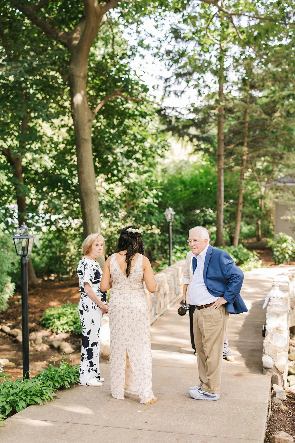 Three people, two women and one man, standing on a sidewalk in a lush green outdoor area, engaged in conversation during daytime.