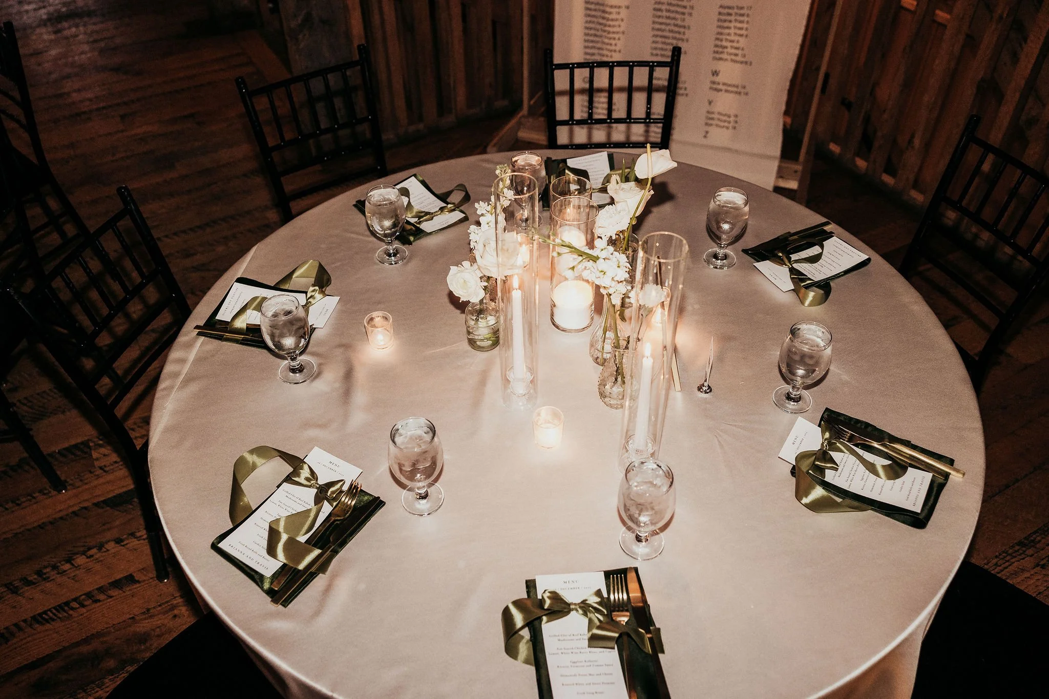 A round table set for a formal event with a beige tablecloth, decorated with white flowers and tall candles in glass holders. Black chairs are arranged around the table, with place settings including black napkins with gold ribbons, silverware, menu 