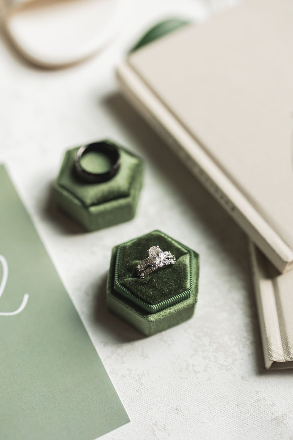 A diamond engagement ring on a green velvet ring box, displayed on a white surface near a book and a smaller green velvet box.