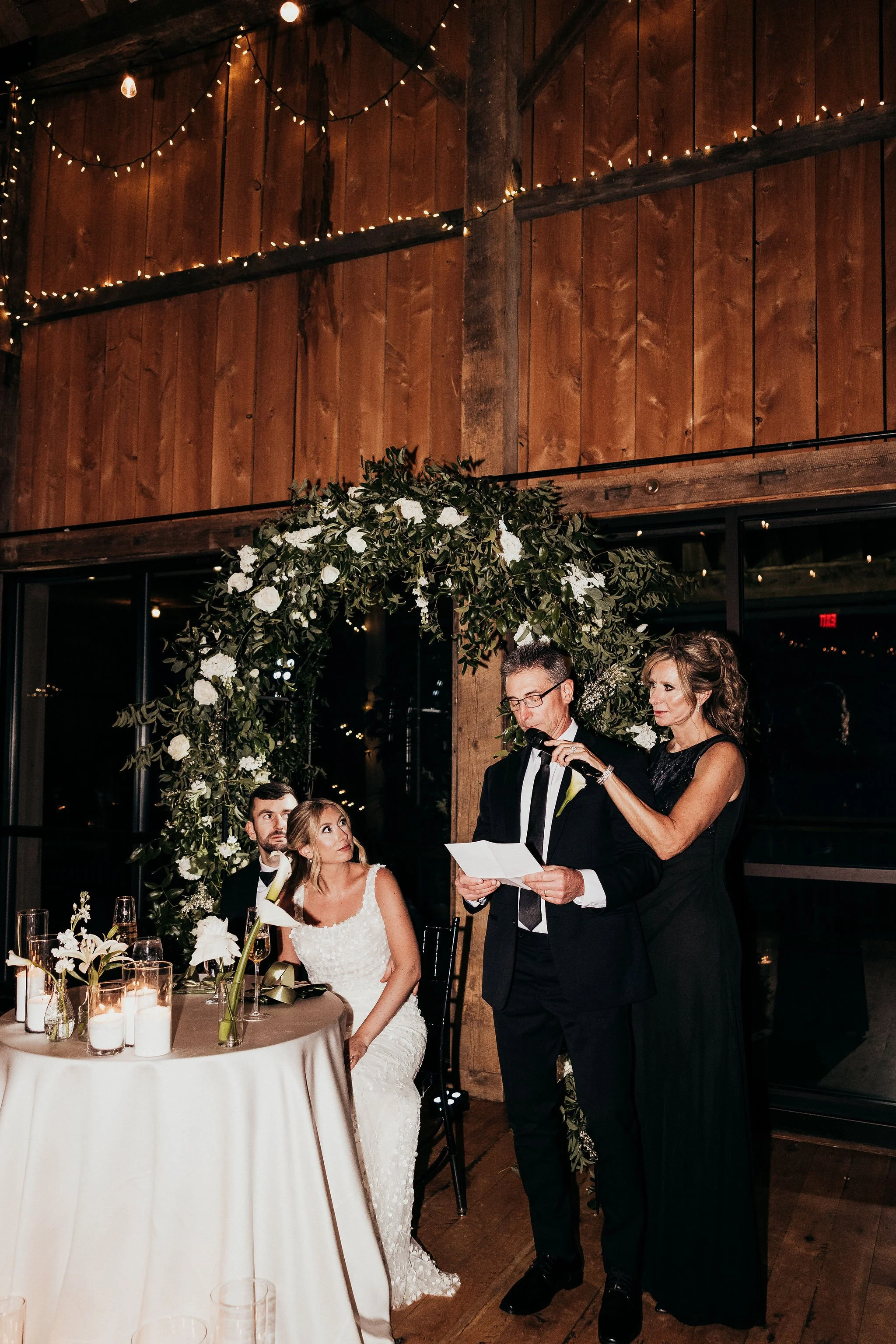 A wedding reception with a bride and groom seated at a table, surrounded by floral arrangements and candles, while an officiant reads vows. Elegant wooden interior with string lights and a floral arch.