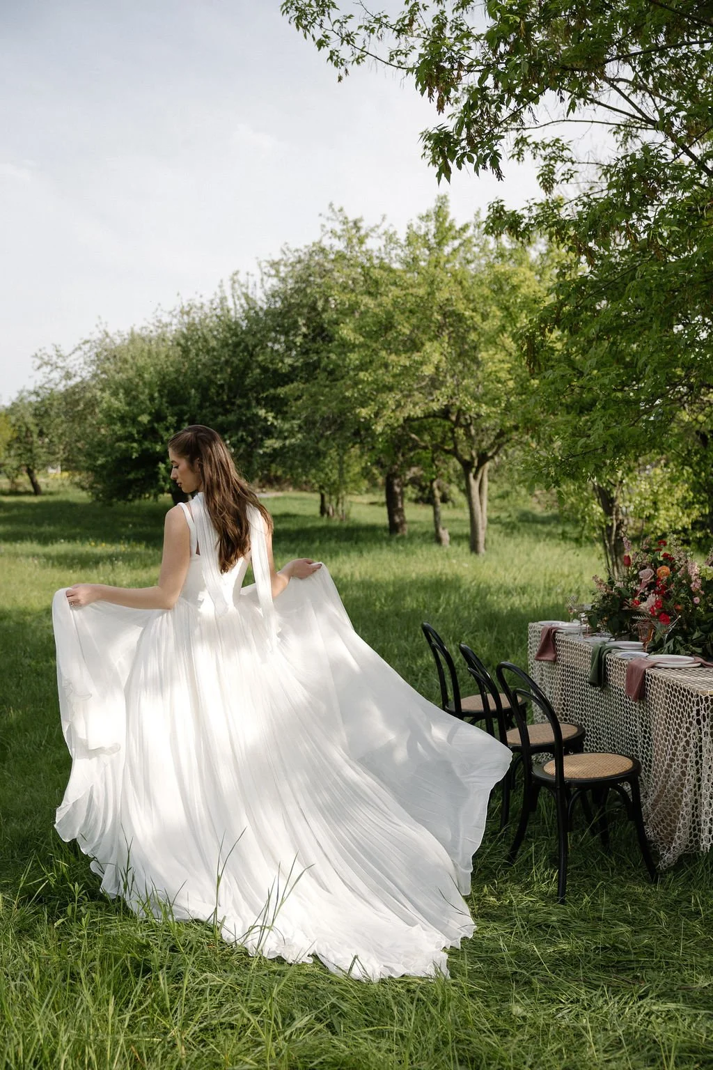 A woman in a flowing white dress holding the sides of her dress in an outdoor setting with green grass and trees, beside a table with chairs and a floral arrangement.