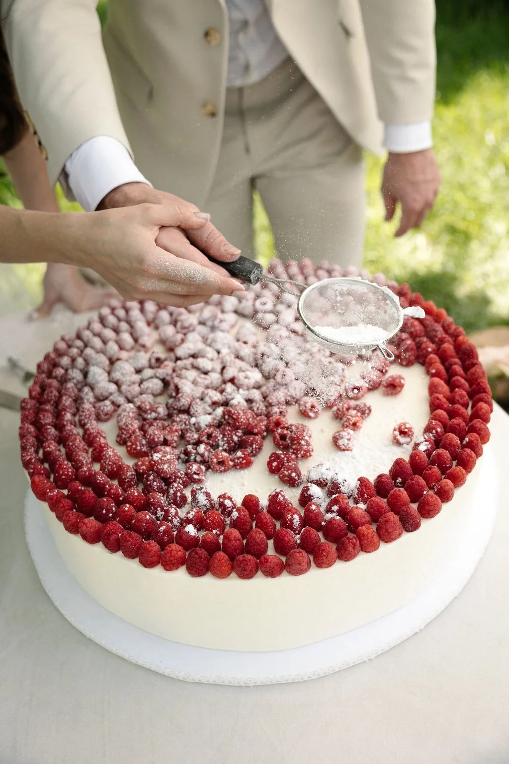Close-up of a wedding cake decorated with red berries, with a person sprinkling powdered sugar on top while another person stands nearby. The scene is outdoors with blurred greenery in the background.