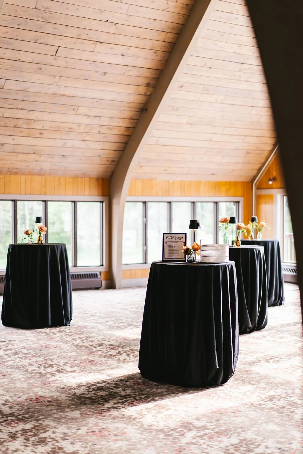 Event space with three round tables covered in black tablecloths, decorated with small flower arrangements, lamps, and a raffle sign, set in a room with wooden walls and large windows, under a vaulted wooden ceiling.