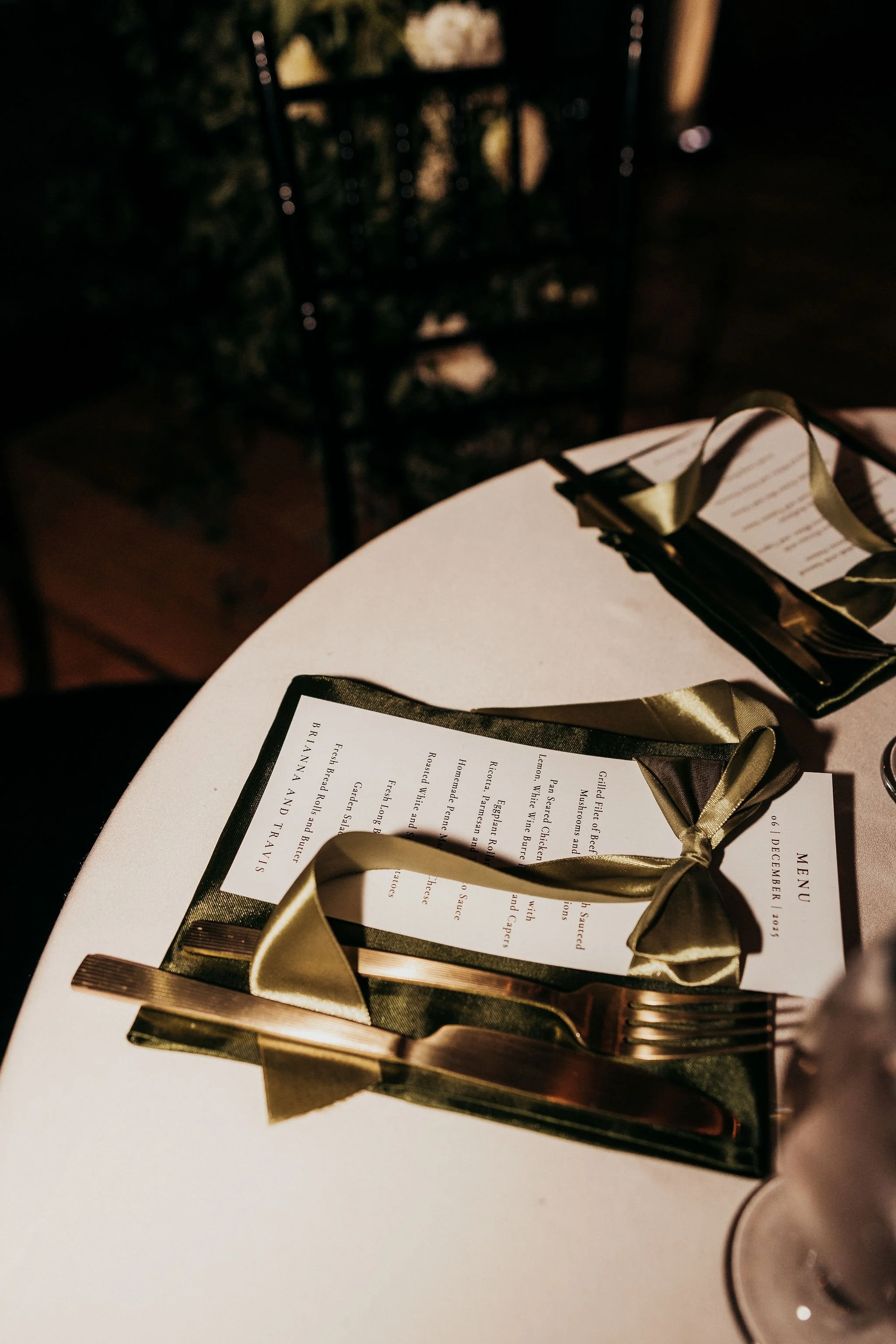A table with two menus tied with golden ribbons, a fork, and a knife. In the background, a blurred wine rack is visible.