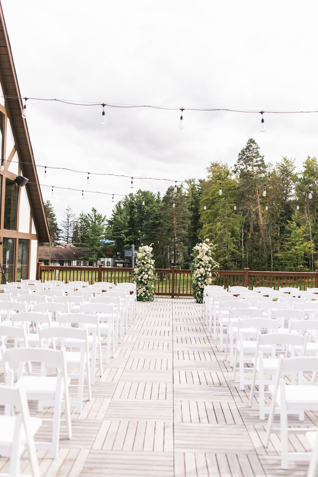 Outdoor wedding ceremony setup on a wooden deck with white chairs, floral arrangements, string lights, surrounded by trees on a cloudy day.