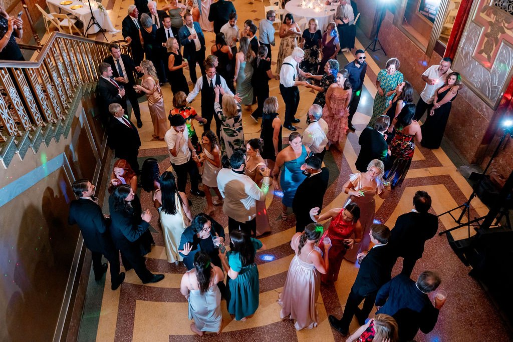 People dancing and socializing at a formal event or party in a spacious hall with ornate walls and wooden floors.