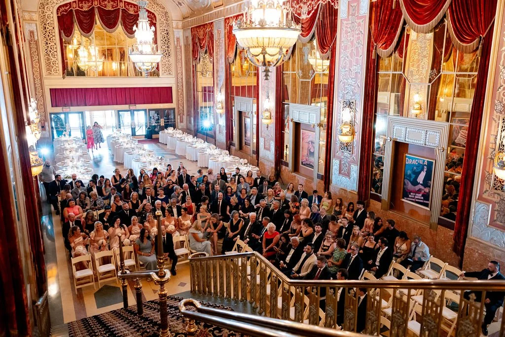 Audience seated in an ornate theater or ballroom with patterned walls, large chandelier, and velvet curtains, attending a performance or event.